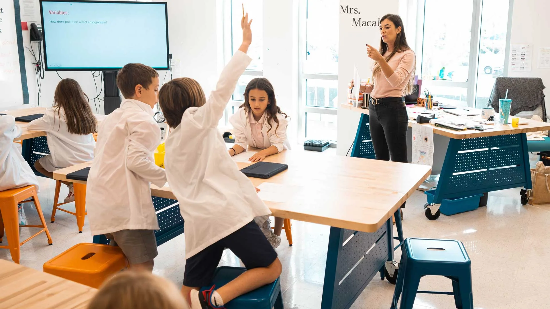 student raising hand while teacher stands instructing the classroom